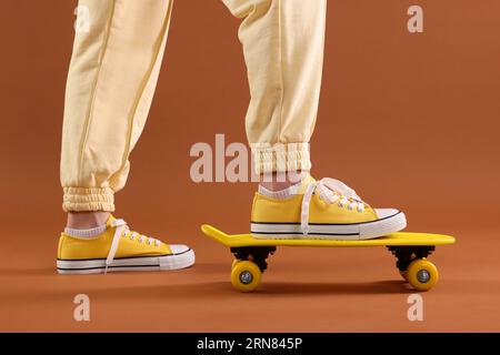Donna in classica sneaker gialla in piedi su penny board su sfondo marrone, primo piano Foto Stock