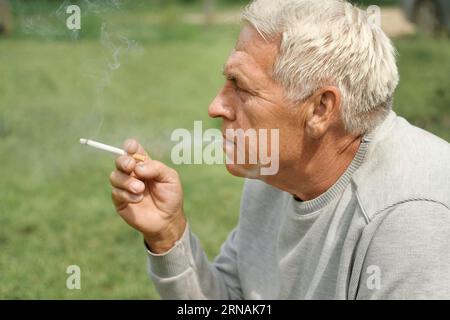 Uomo anziano che accende una sigaretta all'aperto su sfondo verde. Vecchio uomo premuroso che fuma, guardando da parte. Faccia da vicino. Pensione rilassata Foto Stock
