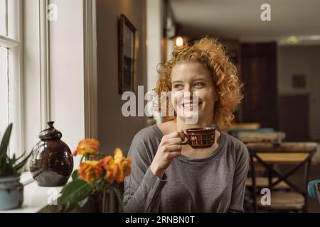 Cheerful young woman sitting at table with cup of coffee in cafe Foto Stock