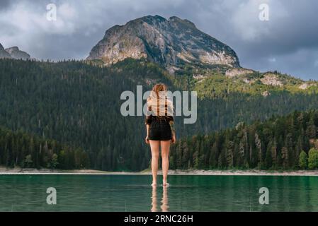 Femmina con lunghi capelli volanti in piedi sull'acqua e vista del lago Nero a Durmitor, Montenegro. La donna che indossa una camicia a quadri ammira il panorama di Foto Stock