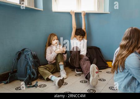 Ragazza che usa lo smartphone seduta da un amico maschio che si allunga mentre è seduta a scuola Foto Stock