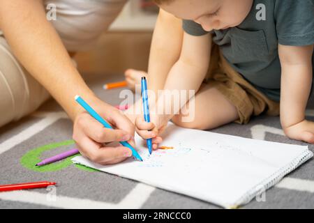 Un bellissimo bambino di due o tre anni con papà disegna con dei marcatori nell'album nella stanza dei bambini di casa seduto sul pavimento. Foto Stock