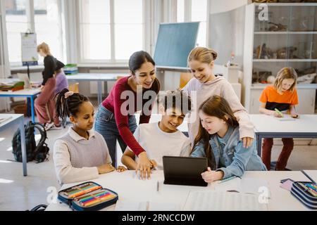 Insegnante con studenti di sesso maschile e femminile che utilizza un tablet PC alla scrivania in classe Foto Stock