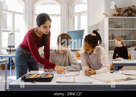 Insegnante che spiega studenti maschi e femmine seduti alla scrivania in classe Foto Stock