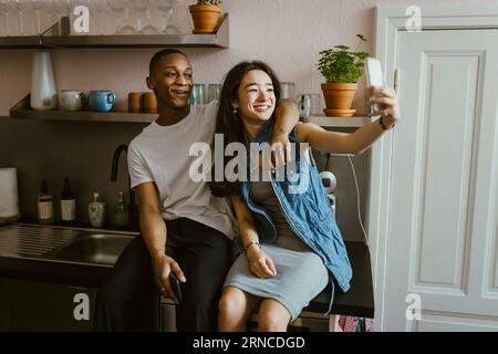 Giovane donna felice che scatta selfie con un amico seduto sul bancone della cucina a casa Foto Stock