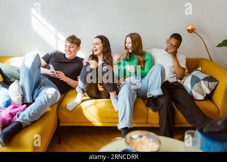 Allegra i giovani amici multirazziali che usano lo smartphone mentre si siedono insieme sul divano a casa