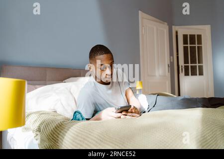 Un giovane che usa uno smartphone mentre è sdraiato a letto a casa Foto Stock
