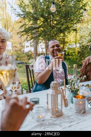 Un uomo gay felice che tiene wineglass seduto con gli amici durante la cena nel cortile posteriore Foto Stock