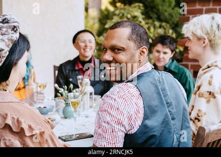 Ritratto di un uomo gay sorridente che guarda a spalla dagli amici durante la cena nel cortile posteriore Foto Stock