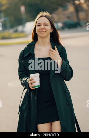 giovane ragazza adolescente con un vestito corto e un lungo impermeabile con un bicchiere di caffè la mattina, la città primaverile, sorridendo guarda la fotocamera Foto Stock