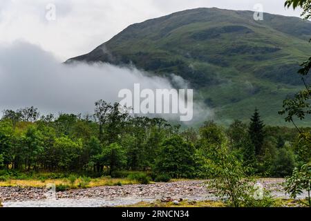 Nuvole e nebbia basse su una foresta con sfondo di montagna (Glencoe, Scozia) Foto Stock