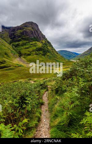 Hiking trail leading into a valley surrounded by steep mountains and a stormy, grey sky Stock Photo