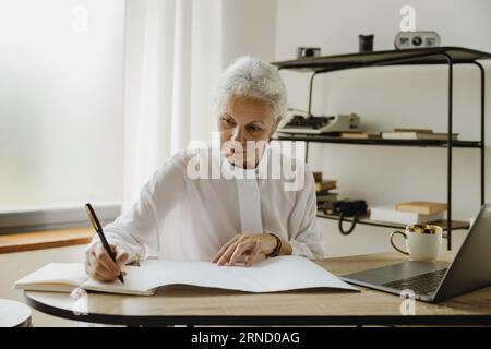 Elegante donna d'affari matura dai capelli grigi seduta alla scrivania con un computer portatile e che prende appunti sul notebook. Foto Stock