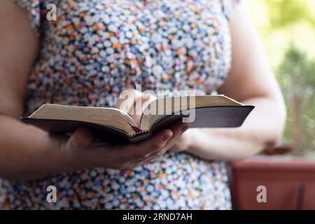 Donna cristiana che legge la sacra bibbia all'aperto. Primo piano. Messa a fuoco selettiva. Persona che tiene un libro con pagine d'oro in mano. Foto Stock