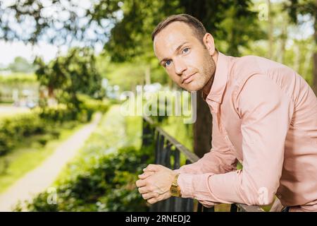 ritratto estivo di un giovane imprenditore in camicia rosa con le mani piegate e orologio da polso dorato mentre si è all'aperto nel parco e si gode il tempo spensierato e.. Foto Stock