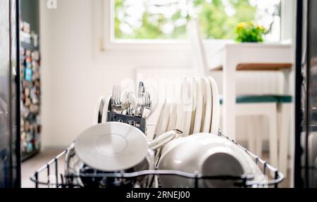 Dish washing machine. Inside an open dishwasher, view to kitchen. Clean plates, utensils, cutlery and pot in washer rack and shelf. Basket with fork. Foto Stock
