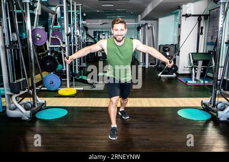 il bell'atleta muscoloso si allena alla funivia con i triceps tirando le maglie con entrambe le braccia e il sorriso soddisfatti con la fotocamera mentre si fa un allenamento duro Foto Stock