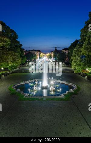 Fontana illuminata con getti d'acqua di fronte al ponte Luitpold e Prinzregenten Street all'ora blu al tramonto a Monaco, Baviera, Germania Foto Stock
