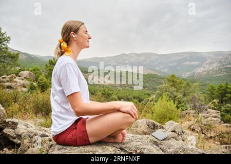 Donna con smartphone seduta in posa di loto su roccia Foto Stock