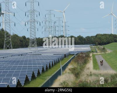 Large solar farm in Vlissingen, Zeeland, the Netherlands with cyclists on bicycle path, wind turbines, and high voltage power lines. Stock Photo