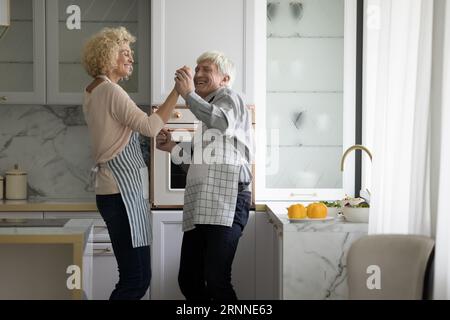 Coppia anziana allegra in grembiuli che ballano con la musica in cucina Foto Stock