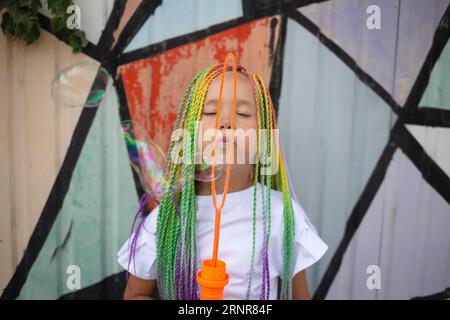 una graziosa bambina con trecce afro multicolore gonfia le bolle di sapone sullo sfondo di una parete splendidamente decorata Foto Stock