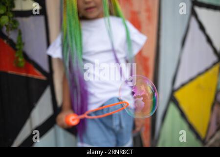 una graziosa bambina con trecce afro multicolore gonfia le bolle di sapone sullo sfondo di una parete splendidamente decorata Foto Stock