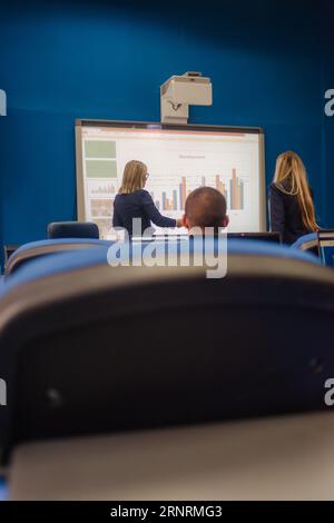 Studentessa bionda con mancanza di conoscenza in piedi accanto al suo professore universitario di fronte a una lavagna intelligente che la ascolta spiegare qualcosa. Foto Stock