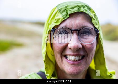 La donna con giacca impermeabile e gocce di pioggia sugli occhiali torna immersa in una passeggiata sul mare, Portbail, Cotentin, Manche, Normandia, Francia Foto Stock