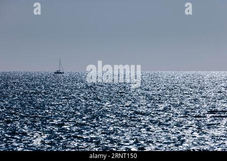 Barca a vela all'orizzonte con il mare scintillante e scintillante contro la luce del Canale della Manica proprio di fronte al tramonto, Portbail, Cotentin Foto Stock