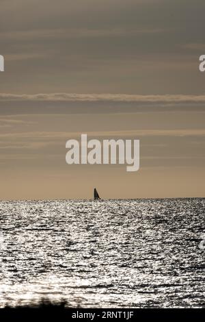 Barca a vela all'orizzonte con il mare scintillante e scintillante contro la luce del Canale della Manica proprio di fronte al tramonto, Portbail, Cotentin Foto Stock