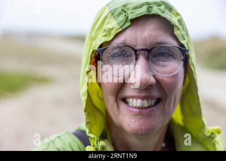 La donna con giacca impermeabile e gocce di pioggia sugli occhiali torna immersa in una passeggiata sul mare, Portbail, Cotentin, Manche, Normandia, Francia Foto Stock