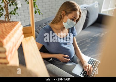 Donna d'affari incinta con maschera medica seduto divano lavoro portatile Foto Stock