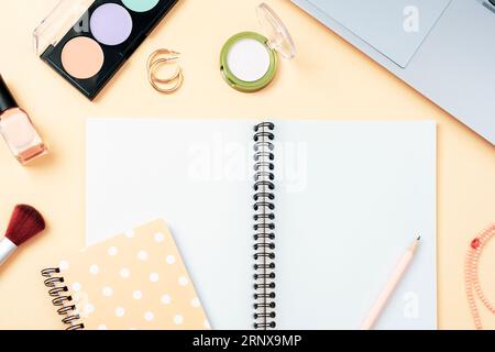 Blocco note vuoto, cosmetici per il trucco e computer portatile su sfondo arancione pastello, vista dall'alto. Design piatto creativo, mockup. Foto Stock