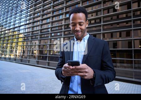 Giovane adulto latino maschio fuori dal moderno edificio degli uffici usando il telefono. Uomo d'affari in abiti formali. Foto Stock