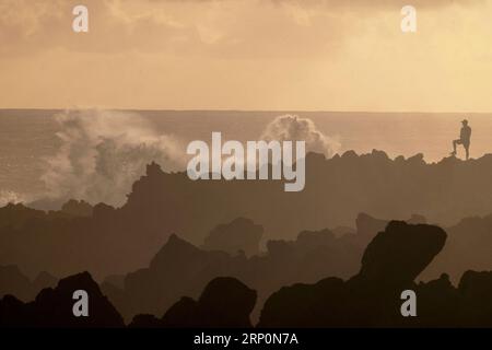 Un escursionista solitario si erge su rocce vulcaniche lungo la costa dell'isola di Réunion, catturata in silhouette contro l'orizzonte dell'oceano e il suggestivo sk tropicale Foto Stock