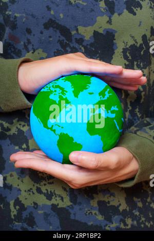 Una donna che indossa abiti mimetici tiene in mano un globo verdeggiante. Concetto di dedizione ambientale creativa. Foto Stock