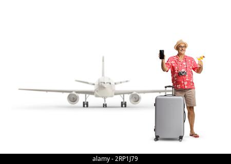 Un turista maturo con una valigia che regge un cocktail e mostra uno smartphone davanti a un aereo isolato su sfondo bianco Foto Stock