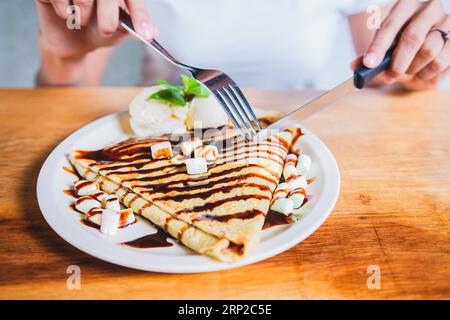 Mani di persona con forchetta che taglia crepe al cioccolato e gelato. Donna che mangia crepe al cioccolato e gelato con la forchetta Foto Stock