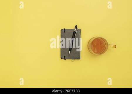 Un blocco note nero con una penna e una tazza di tè su una superficie gialla Foto Stock