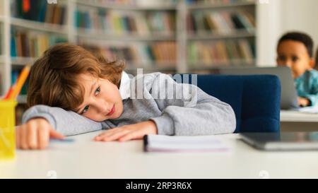 Studente annoiato sdraiato sul tavolo in biblioteca o in classe durante una lezione non interessata, guardando lontano e pensando Foto Stock