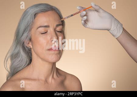 Concetto di sollevamento della pelle. Colpo alla testa di una donna anziana che riceve un'iniezione di filler di bellezza sulla fronte, sfondo beige Foto Stock