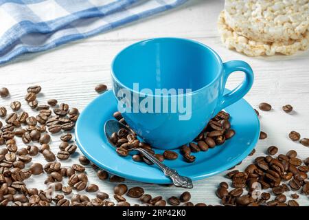 Caffè mattutino - una tazza blu vuota con un piattino per il caffè e chicchi di caffè sparsi e torte di riso su un tavolo di legno bianco. Foto Stock