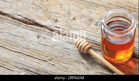 Un vaso di vetro con miele e un bastoncino su fondo di legno. Recipiente trasparente con prodotto nutrizionale color oro. Sopra la vista, spazio di copia Foto Stock