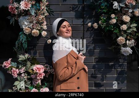 Ritratto di una giovane donna caucasica vestita in un hijab vicino a un muro con fiori. Foto Stock