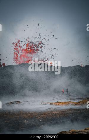 Eruzione vulcanica a Litli hrutur Islanda vicino a fagradalsfjall 2023. 2 persone stanno scattando foto di lava che esplode sul bordo di un cratere. Lava nera. Foto Stock