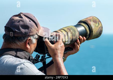 uomo anziano che indossa un berretto da baseball che utilizza un grande teleobiettivo su una fotocamera reflex digitale. uomo di mezza età che indossa un cappello con una grande fotocamera. Foto Stock