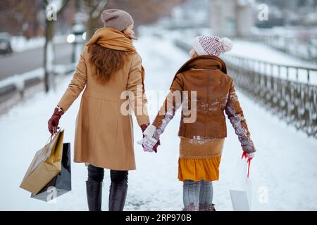 Vista da dietro la mamma e il bambino eleganti in cappotto, cappello, sciarpa e guanti con borse per lo shopping che camminano all'aperto in città in inverno. Foto Stock