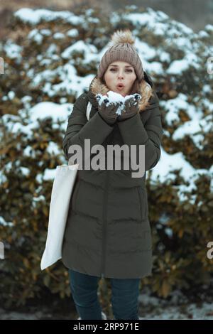 donna moderna con cappotto verde e cappello marrone all'aperto nel parco cittadino in inverno con guanti e berretto vicino a rami innevati. Foto Stock