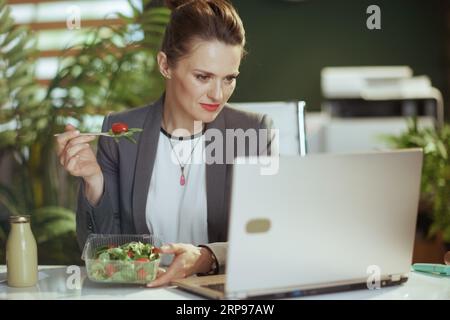 Ambiente di lavoro sostenibile. moderna proprietaria di una piccola impresa di 40 anni, in abito da lavoro grigio, in un moderno ufficio verde con un laptop che mangia insalata. Foto Stock